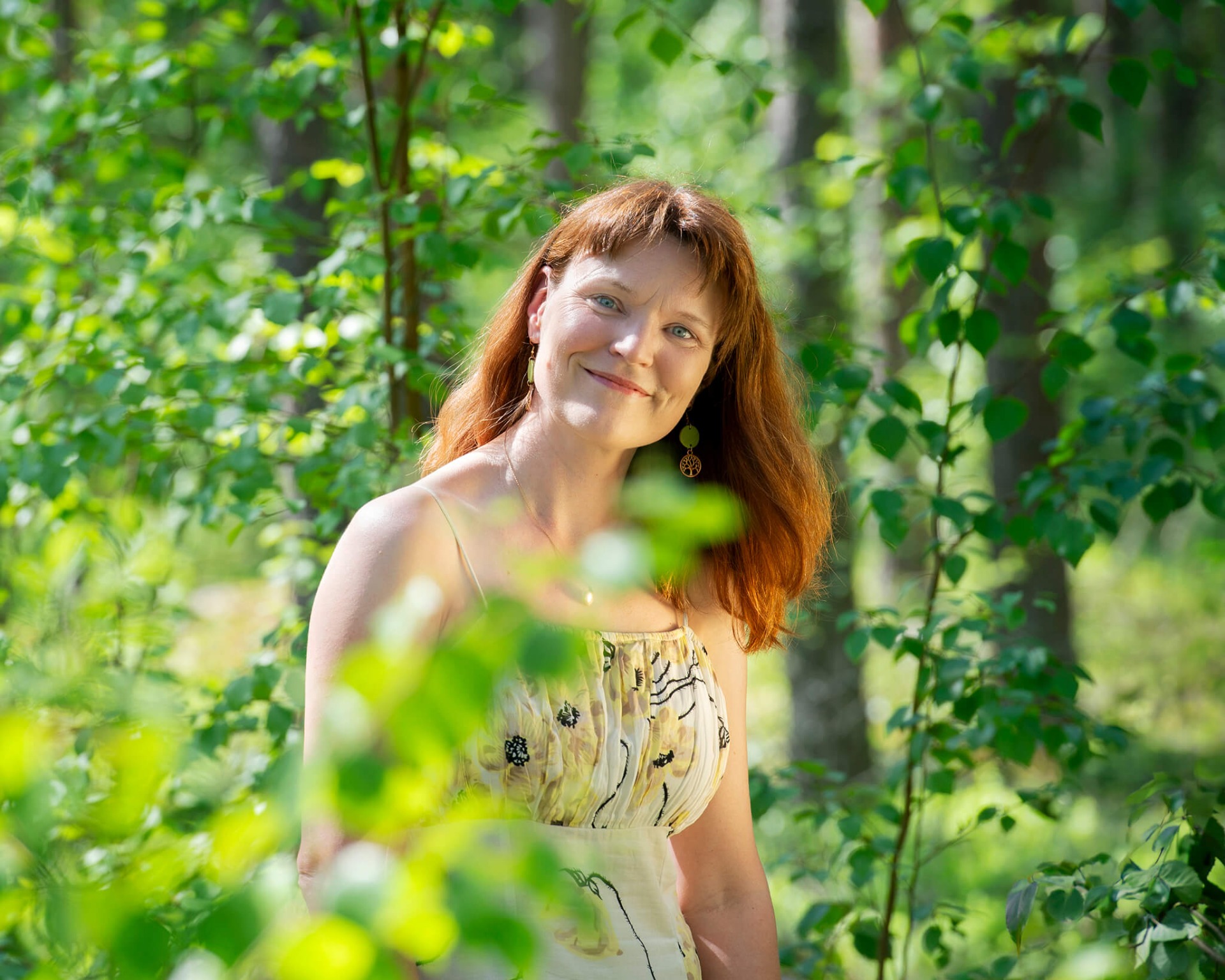 Punatukkainen nainen hymyilee metsässä, vihreiden lehtien ja pehmeän taustan keskellä.